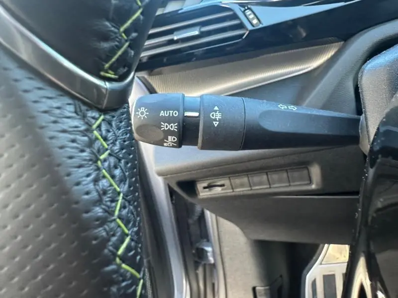 Gros plan sur la commande des phares à gauche du volant cuir noir avec surpiqûres vertes, intérieur gris du Peugeot 308.