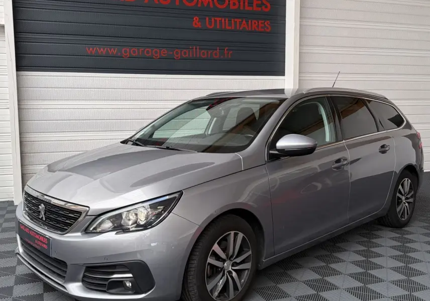 Peugeot 308 BlueHDi 130 S&S Allure gris foncé vue 3/4 avant droit, avec jantes alliage et phares LED allumés.