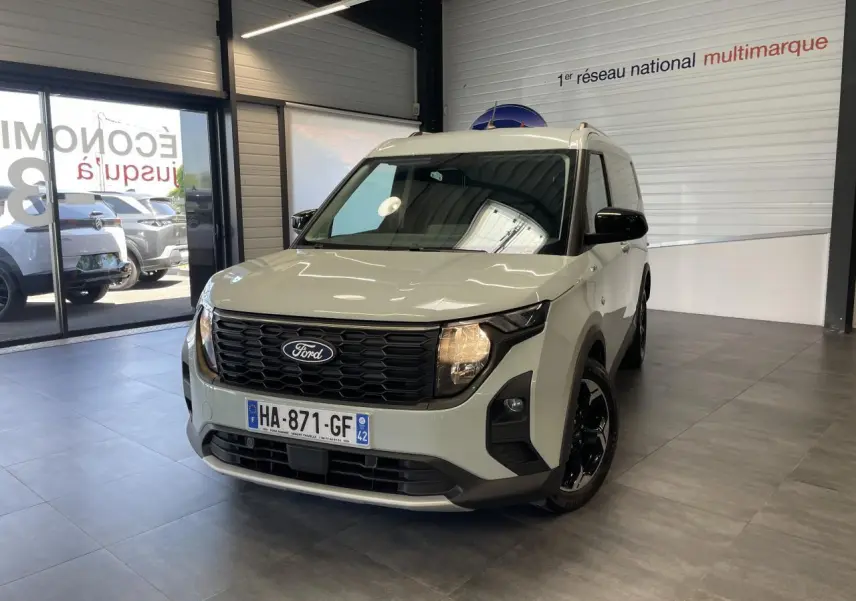 Ford Tourneo Courier gris clair vu en 3/4 avant droit, avec calandre noire nid d'abeille et jantes noires brillantes.