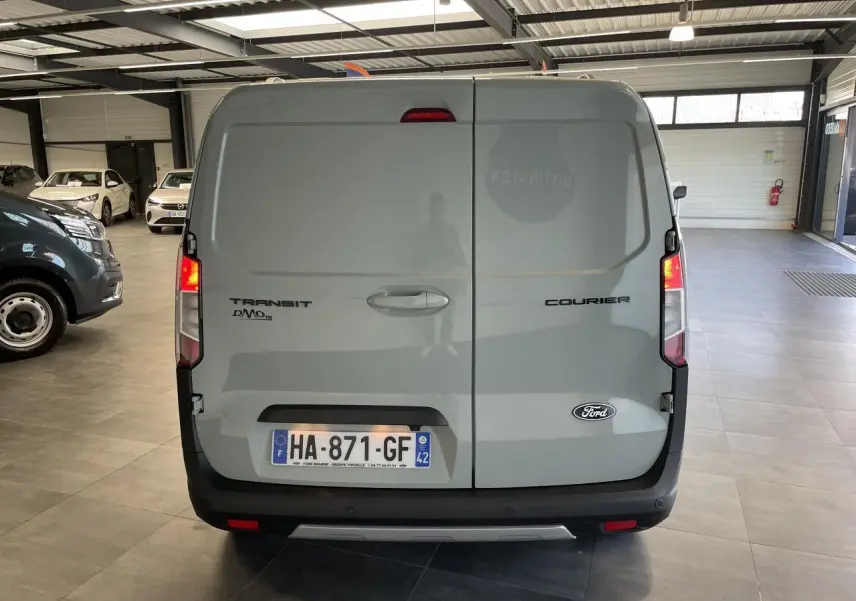 Vue arrière d'un Ford Tourneo Courier gris clair avec portes battantes et feux arrière allumés en intérieur showroom.