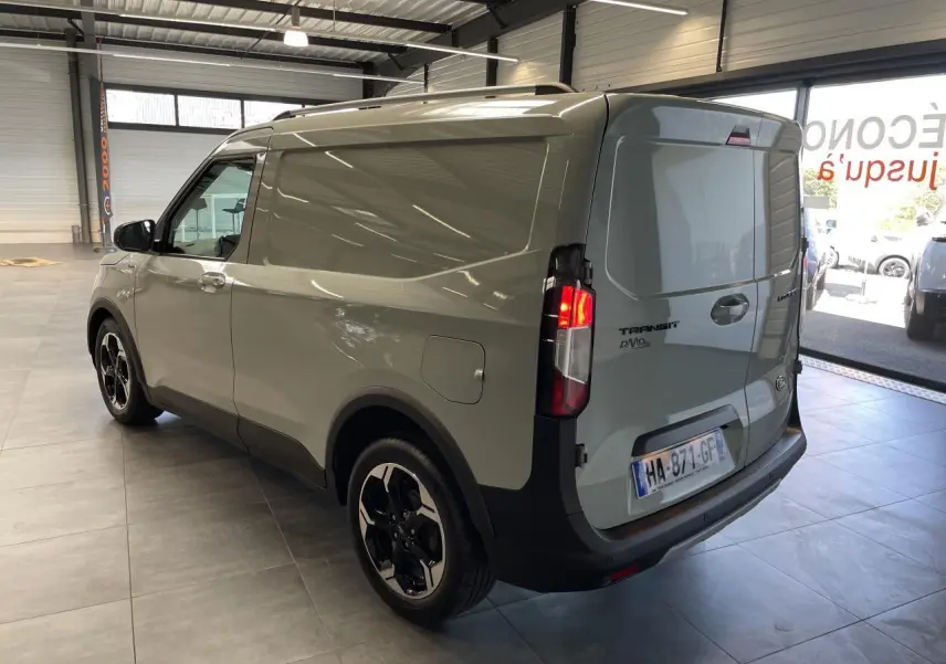 Vue 3/4 arrière droite du Ford Tourneo Courier gris clair avec jantes aluminium et feux arrière allumés en intérieur showroom.
