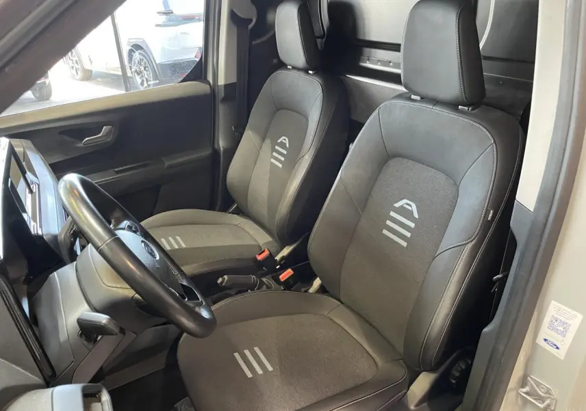 Vue intérieure côté conducteur du Ford Tourneo Courier gris clair 2024, sièges tissu avec logo et volant cuir synthétique.