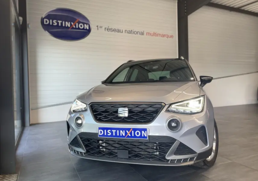 Vue avant d'un SEAT Arona 2024 gris clair avec calandre noire et phares allumés en intérieur showroom.