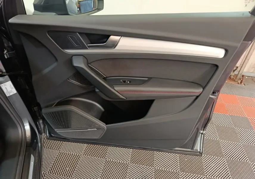 Vue rapprochée de la porte avant gauche d’un Audi Q5 gris clair 2023, avec garniture noire et surpiqûres rouges.