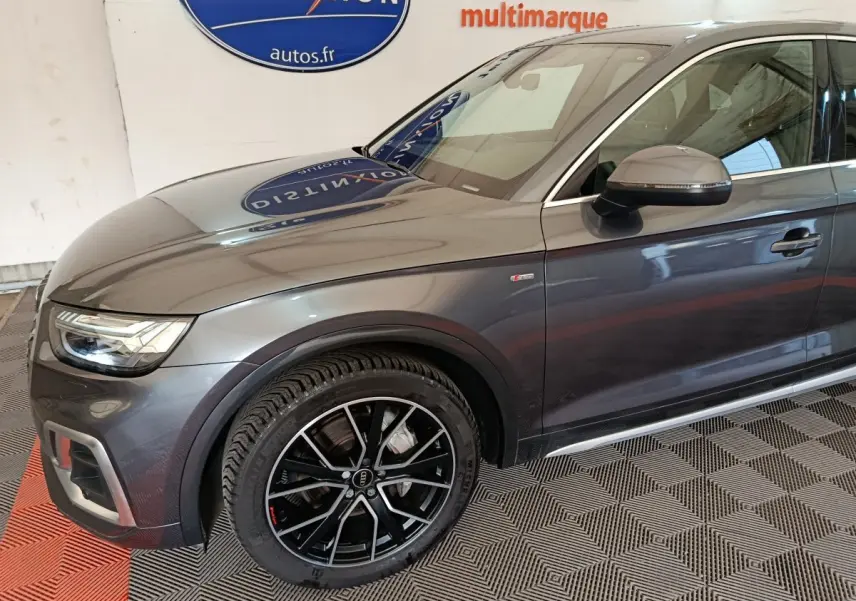 Vue de profil avant droit d'un Audi Q5 gris clair 2023 avec jantes noires et badge S line sur l'aile avant.