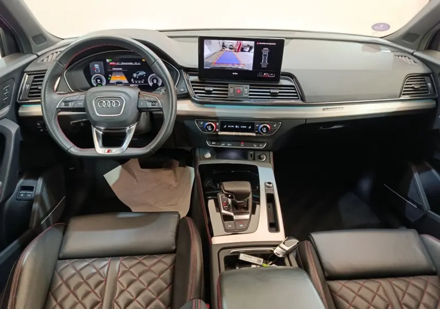 Vue intérieure avant de l'Audi Q5 2023 hybride rechargeable, avec tableau de bord moderne et sièges en cuir noir surpiqués rouges.