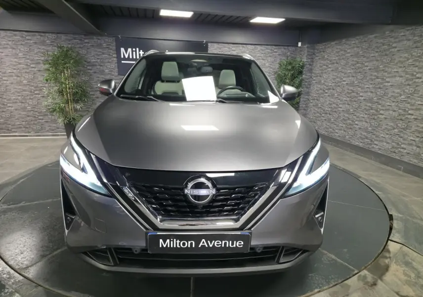 Vue avant d'un Nissan Qashqai 1.5 e-Power gris métallisé avec phares LED allumés en intérieur showroom.