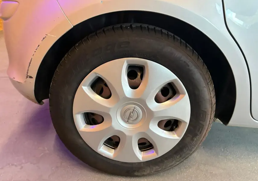 Gros plan sur la roue arrière droite d’une Opel Corsa gris clair de 2012 avec enjoliveur d’origine et carrosserie légèrement éraflée.