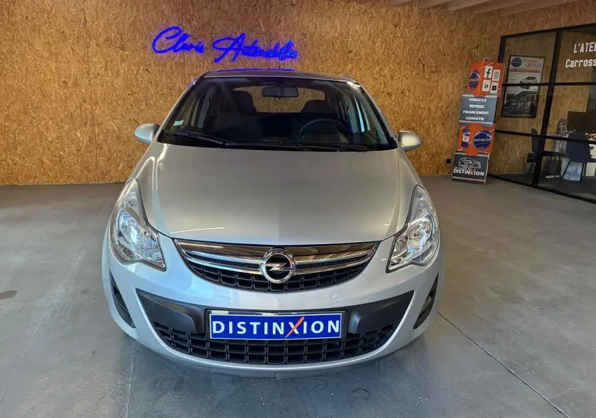 Vue de face d'une Opel Corsa gris clair 2012, avec calandre chromée et plaque Distinxion, en intérieur showroom.