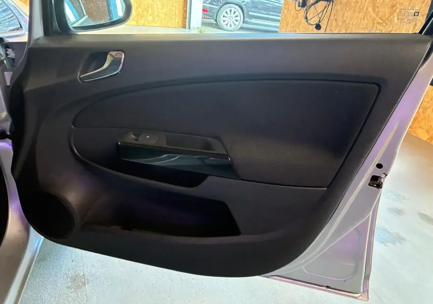 Vue intérieure de la porte avant gauche d’une Opel Corsa gris clair 2012 avec garniture noire et poignée argentée.