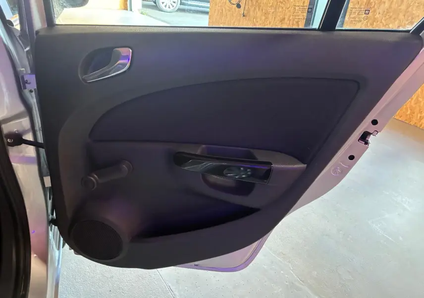 Vue rapprochée de la porte arrière gauche ouverte d’une Opel Corsa gris clair, avec poignée intérieure noire et manivelle de vitre.