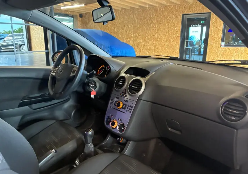 Vue intérieure côté conducteur de l’Opel Corsa gris clair 2012, tableau de bord et volant avec levier de vitesse manuel visibles.