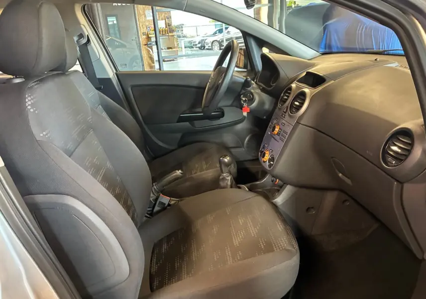 Vue intérieure côté conducteur de l’Opel Corsa 2012, sièges tissu noir et tableau de bord avec commandes climatisation.
