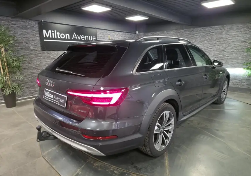 Audi A4 Allroad gris vue 3/4 arrière droit avec feux allumés et crochet d'attelage visible en intérieur showroom.
