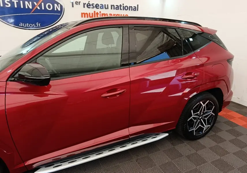 Vue latérale gauche du Hyundai Tucson rouge 2022 avec jantes noires et marchepieds argentés visibles en intérieur showroom.