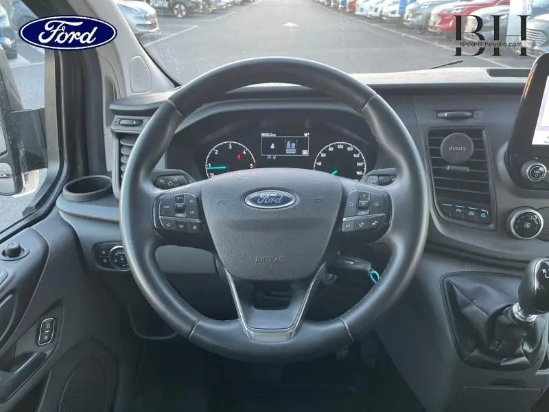 Vue centrée sur le volant cuir et tableau de bord du Ford Transit Custom gris, avec levier de vitesses manuel visible à droite.