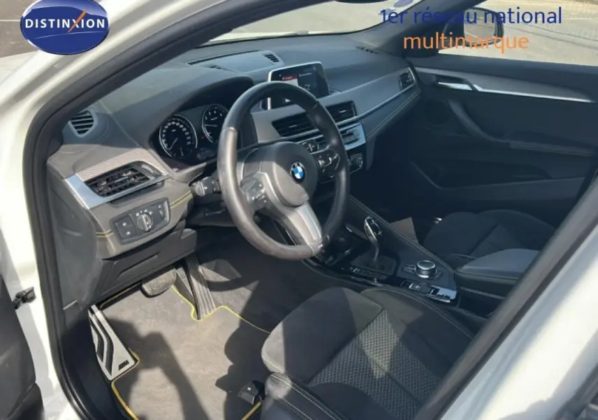 Intérieur côté conducteur de BMW X2 blanc, volant sport, console centrale avec boîte auto et écran GPS visible.