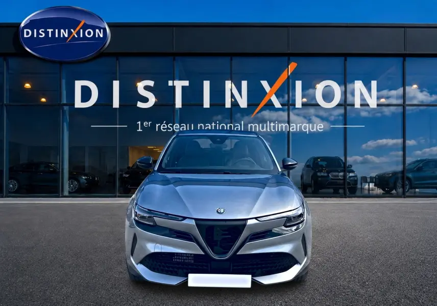 Vue frontale d'une Alfa Romeo Junior 1.2 hybride gris métallisé devant un showroom Distinxion avec reflets de ciel bleu.