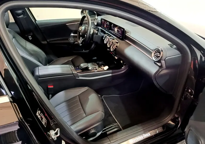 Vue intérieure côté conducteur d'une Mercedes Classe A berline noire, avec tableau de bord digital et aérateurs ronds chromés.