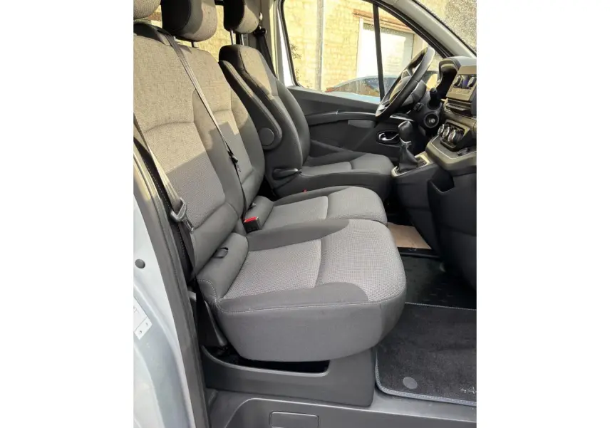 Vue latérale intérieure côté conducteur du Renault Trafic Fourgon gris clair, montrant les sièges avant et le tableau de bord.