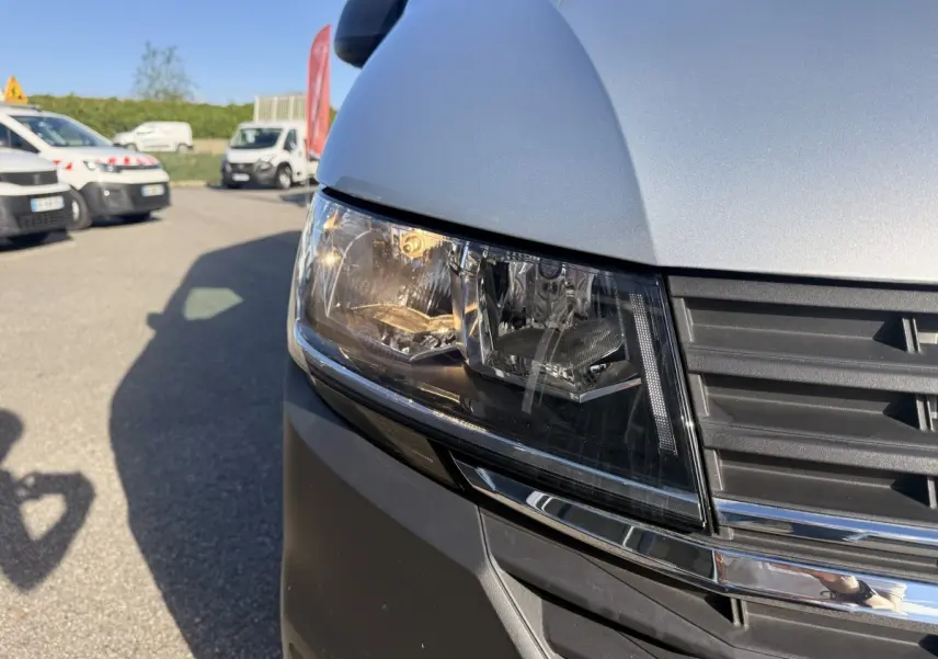 Gros plan sur le phare avant droit et la calandre d’un Volkswagen Transporter gris clair sous un ciel bleu.