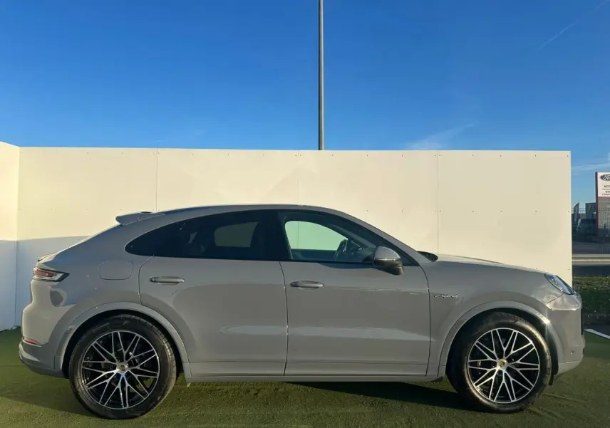 Vue de profil côté gauche d'un Porsche Cayenne Coupé 2024 gris arctique avec jantes noires et badge hybride visible.