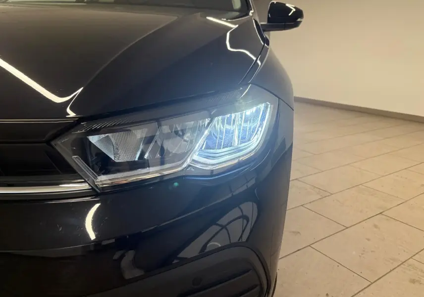 Gros plan sur le phare avant droit allumé d'une Volkswagen Polo noire intense, avec reflets sur la carrosserie.