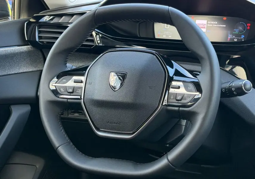 Vue rapprochée du volant multifonction noir de la Peugeot 308 1.2 Hybrid 145ch Allure 2025 avec tableau de bord numérique.