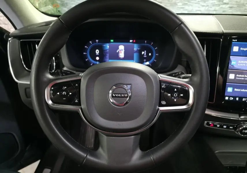 Vue rapprochée du volant noir de la Volvo XC60 2023 avec commandes intégrées et tableau de bord digital en arrière-plan.