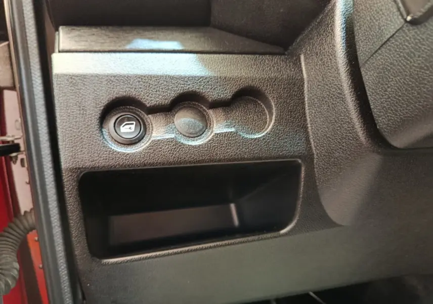Détail intérieur montrant le bouton d'ouverture du coffre sur la partie gauche du tableau de bord noir de l'AIXAM Ambition rouge.