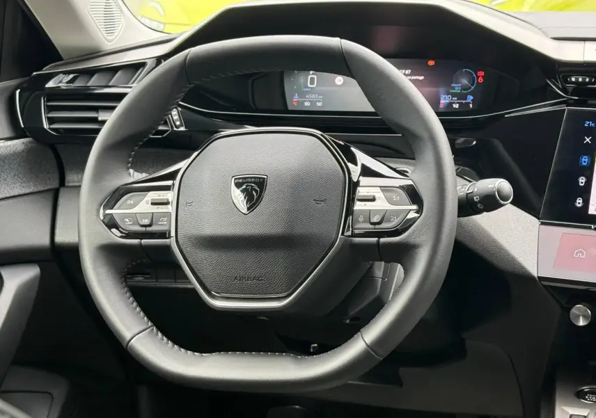 Vue rapprochée du volant multifonction noir de la Peugeot 308 SW 1.2 Hybrid avec tableau de bord numérique en arrière-plan.