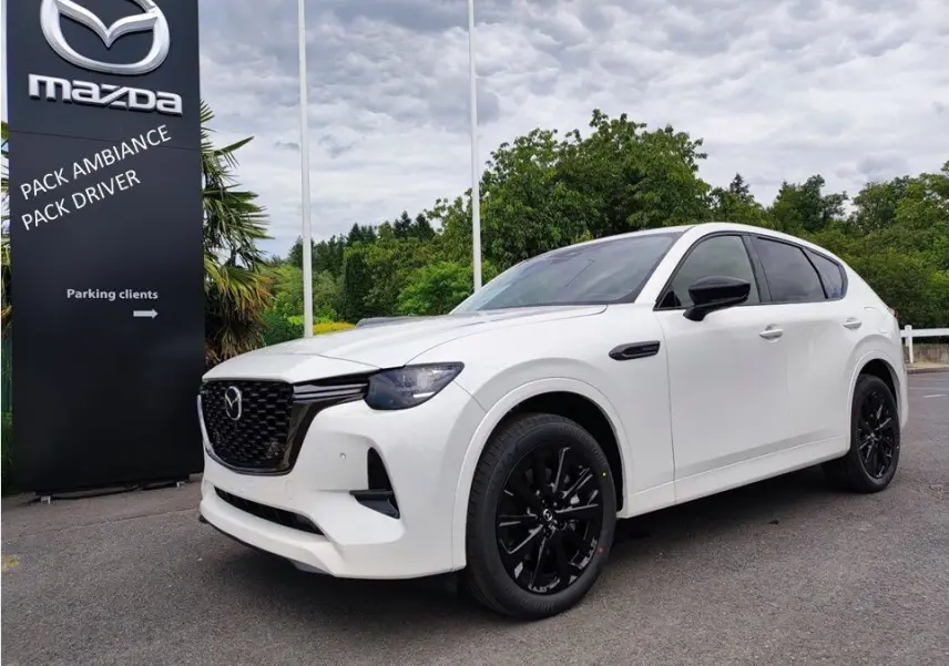 Vue 3/4 avant droite d'un Mazda CX-60 blanc métal avec jantes noires et vitres teintées, devant un panneau Mazda.