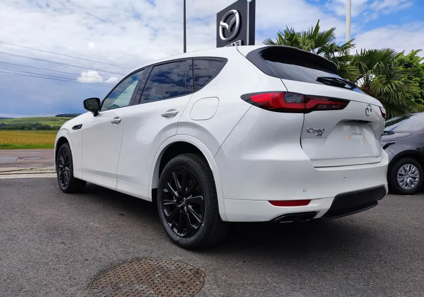 Vue 3/4 arrière gauche d'un Mazda CX-60 blanc métal avec jantes noires et vitres teintées, stationné devant un concessionnaire.