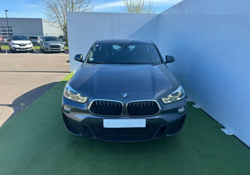 Vue frontale d'un BMW X2 gris foncé avec calandre double haricot et phares LED allumés sur fond urbain.