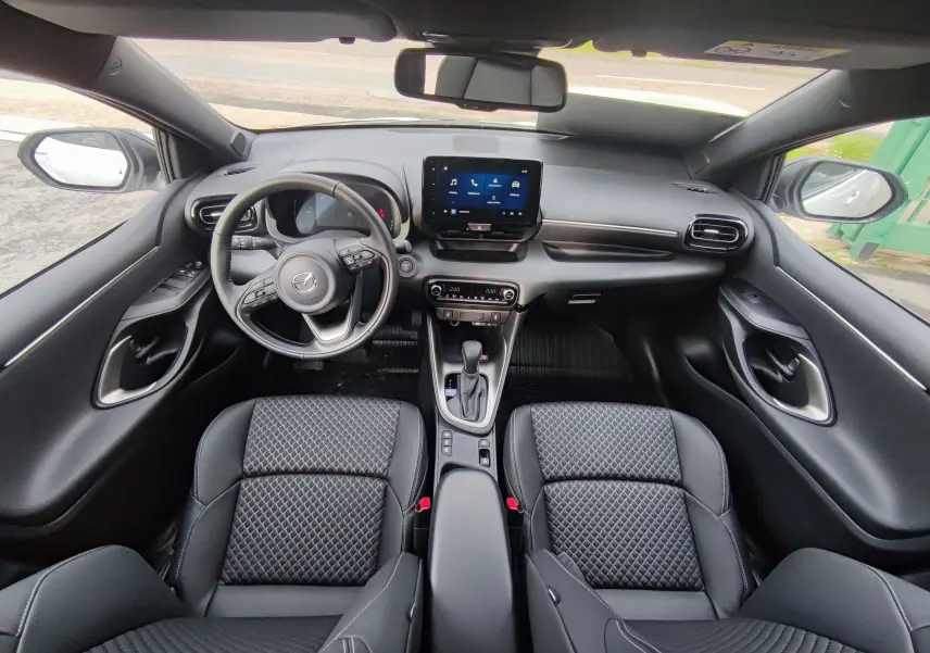 Vue intérieure frontale de la Mazda2 Hybrid 2024 avec sièges noirs matelassés et tableau de bord moderne équipé d'un écran tactile.