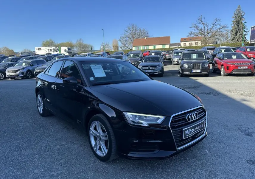 Audi A3 berline noire vue en 3/4 avant droit sur un parking avec plusieurs autres véhicules en arrière-plan.