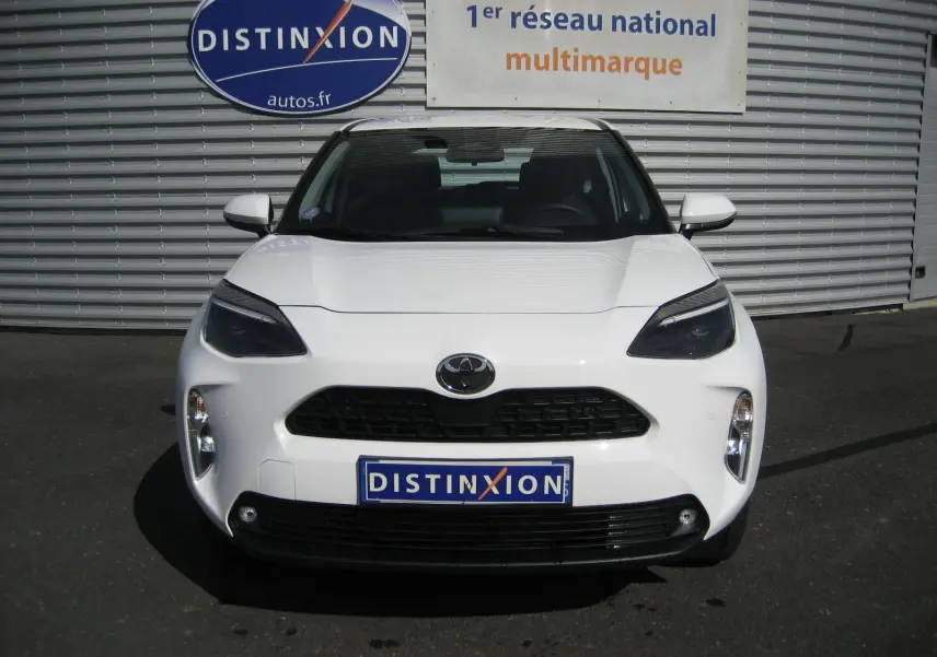 Vue avant d'une Toyota Yaris Cross blanche 2025 avec calandre noire et phares LED sombres, stationnée devant un mur métallique.