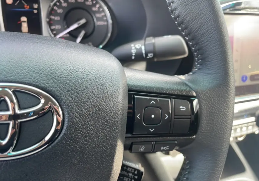 Gros plan sur le volant en cuir noir du Toyota Hilux 2025, avec commandes multifonctions et logo chromé visible.