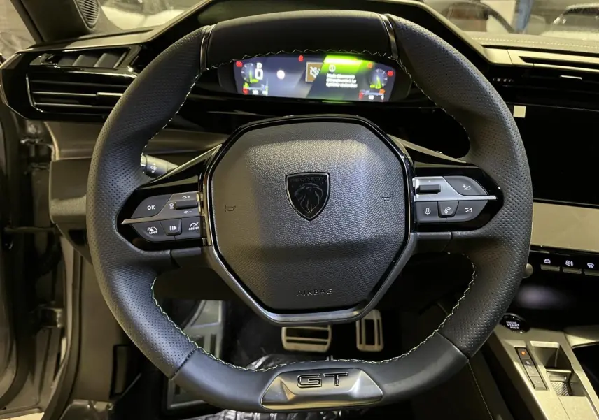 Volant cuir noir GT de la Peugeot 308 2024 vu de face avec combiné numérique et pédaliers aluminium visibles.