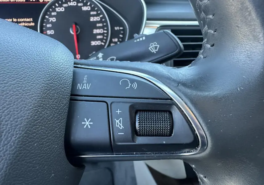 Gros plan sur les commandes vocales et volume au volant cuir noir de l'Audi A6 gris 2015, tableau de bord en arrière-plan.