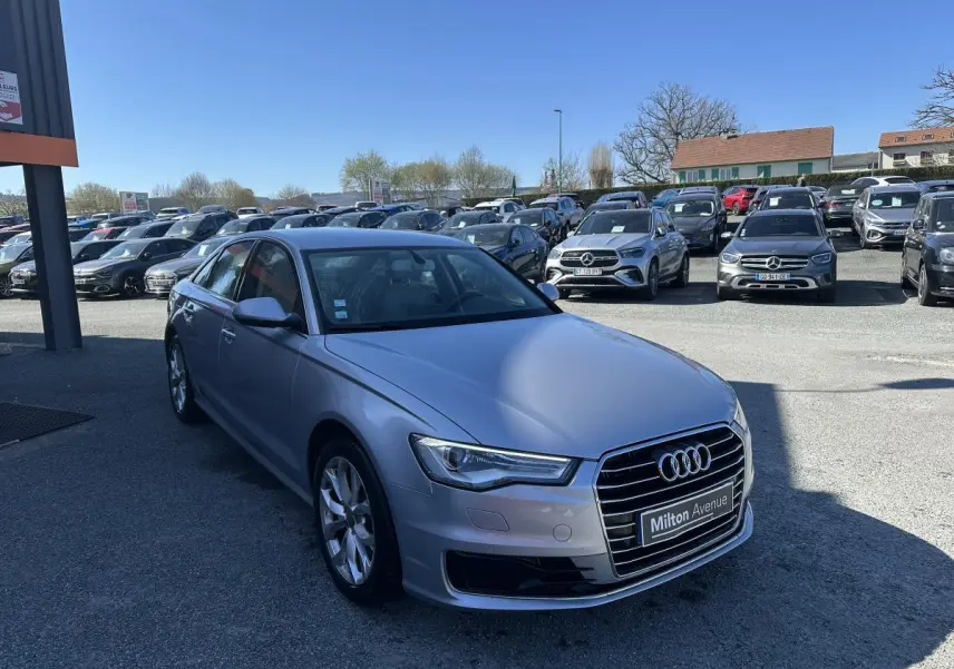 Audi A6 gris vue 3/4 avant droit sur parking avec calandre Singleframe chromée et jantes aluminium.