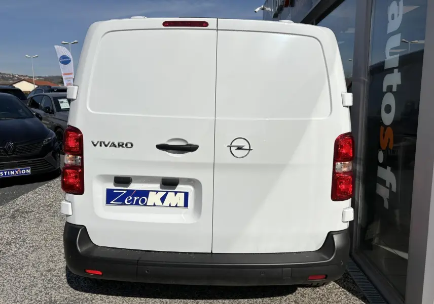Vue arrière d’un utilitaire Opel Vivaro blanc avec portes battantes et pare-chocs noir, stationné près d’un bâtiment vitré.