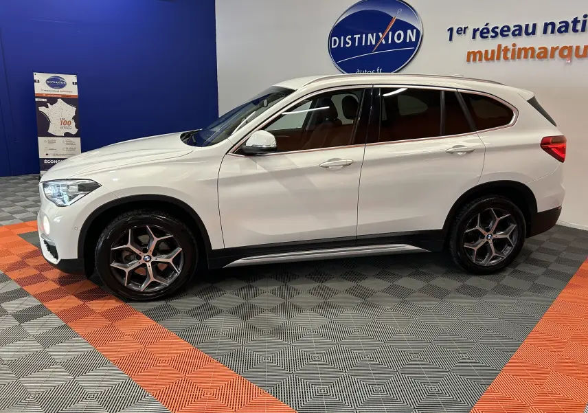 BMW X1 sDrive18iA blanc en vue de profil côté gauche, avec jantes alliage et hayon électrique visible en arrière-plan.