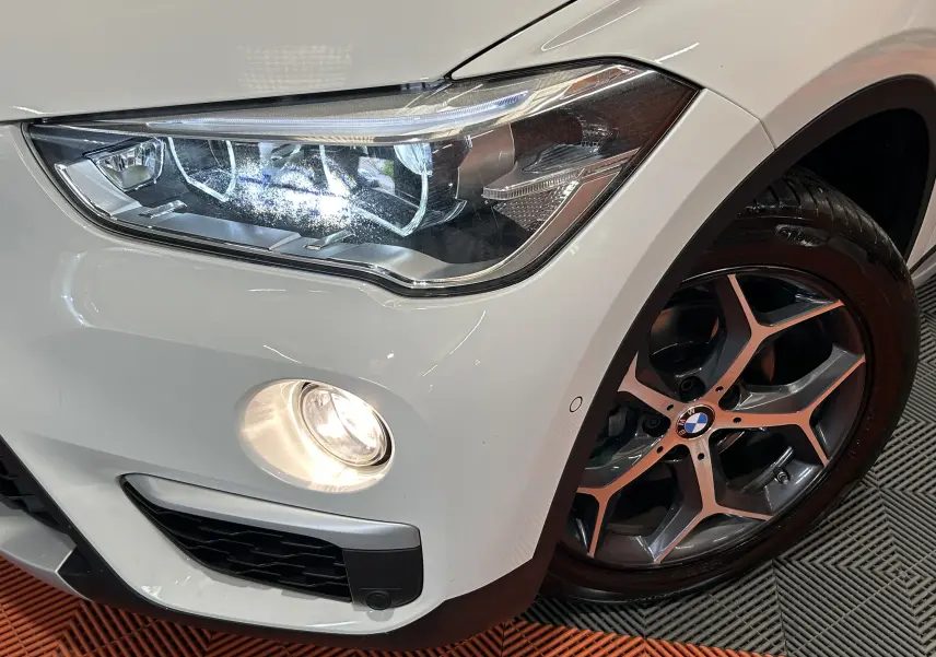 Gros plan sur l'avant droit blanc du BMW X1 sDrive18iA 2018, avec phare LED allumé et jante alliage distinctive.