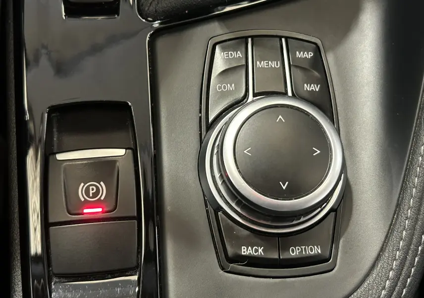 Gros plan sur la molette de commande iDrive et le bouton de frein à main électronique avec témoin rouge, intérieur noir.