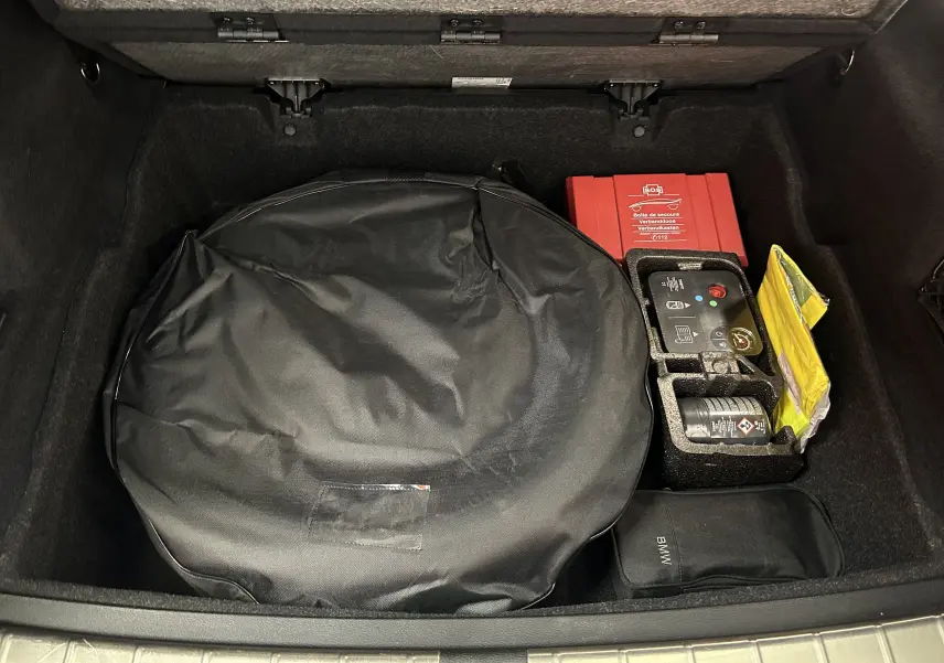 Vue plongeante sur le coffre ouvert d'un BMW X1 blanc, montrant une roue de secours sous housse noire et une trousse de secours rouge.