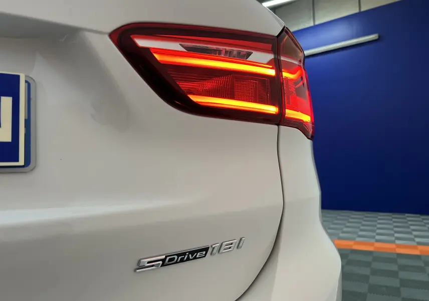 Gros plan sur le feu arrière droit allumé et le badge sDrive18i d’un BMW X1 blanc, arrière droit visible en intérieur.