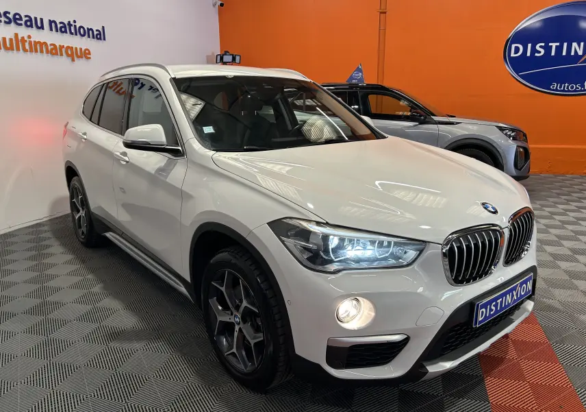 BMW X1 sDrive18iA blanc vue 3/4 avant droit avec phares allumés et jantes foncées en concession.