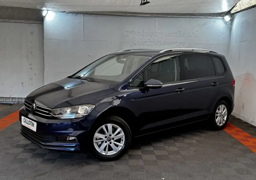 Volkswagen Touran bleu 3/4 avant droit en intérieur, avec jantes alliage et barres de toit chromées.