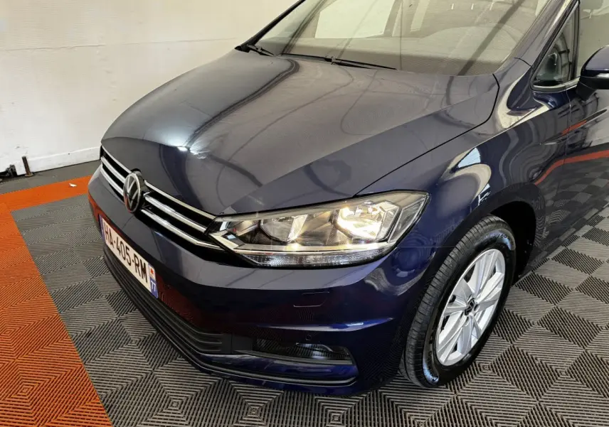 Vue 3/4 avant droit d'un Volkswagen Touran bleu 2024 avec phares allumés sur sol à damier gris et orange.